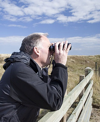 Bild zeigt einen Mann, der mit dem Fernglas in einer Graslandschaft steht und etwas beobachtet.